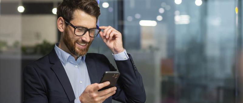 Een lachende man in een net pak en bril kijkt naar zijn smartphone, met een moderne kantoorachtergrond.